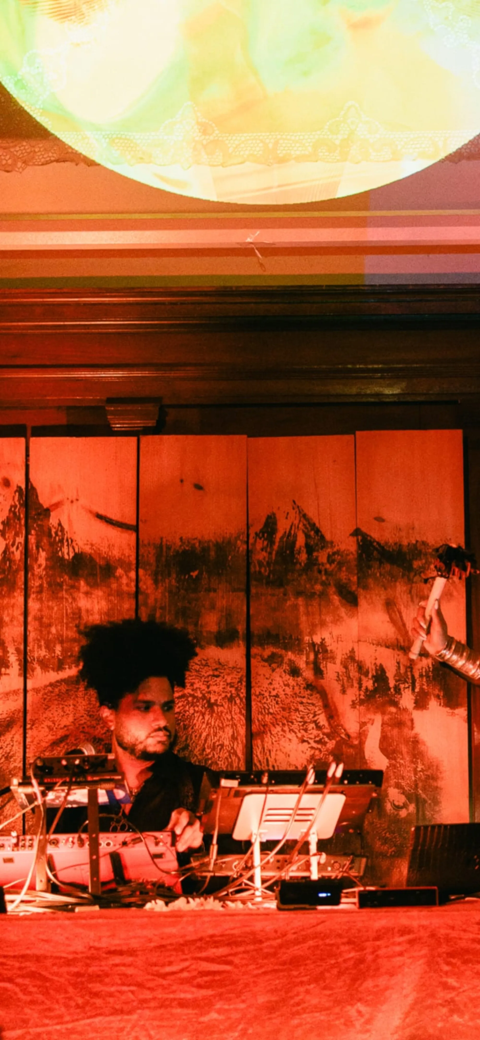 Global artist Benjamin Urbaez and DJ Dreeemy performing with electronic music equipment and projected visuals at live venue with warm red lighting